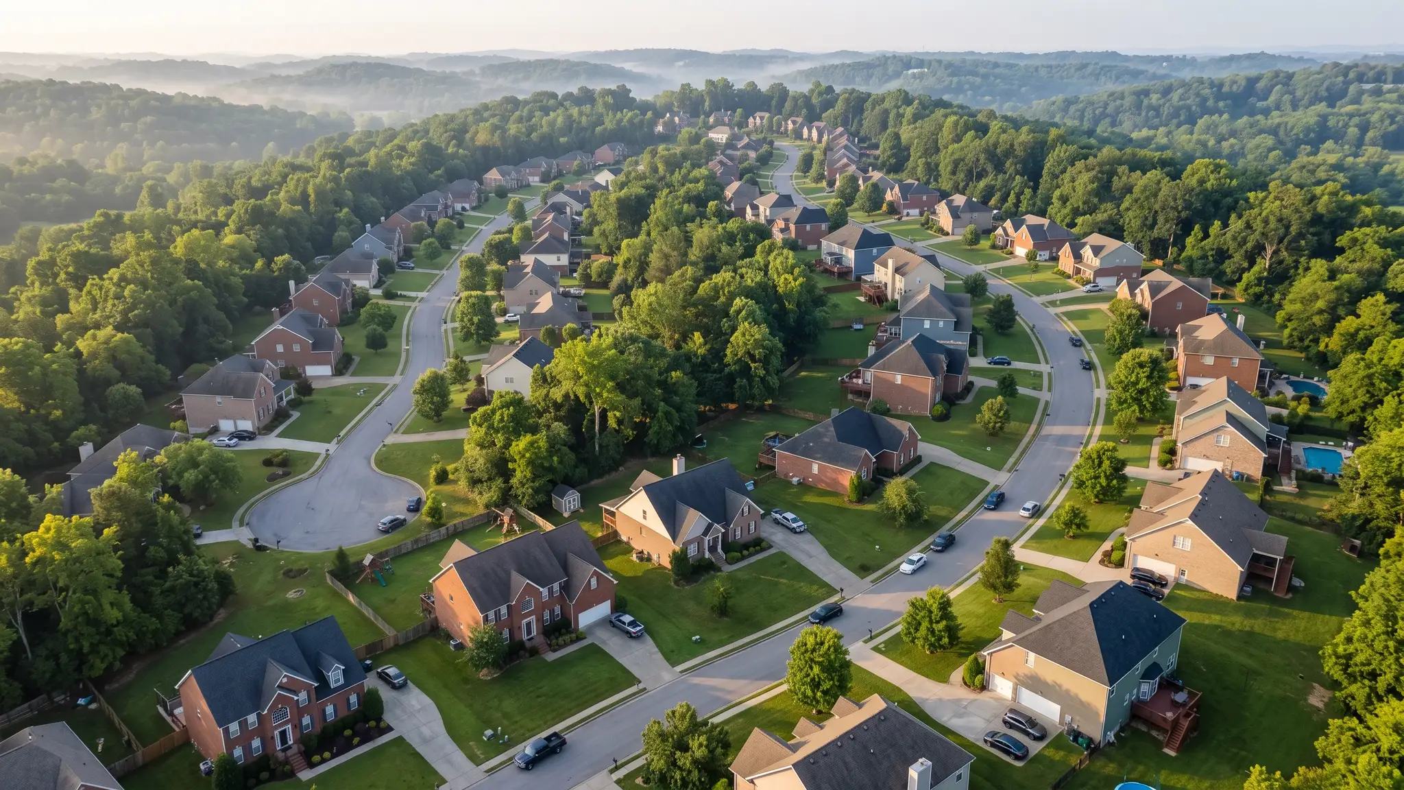 An aerial view of a Tennessee neighborhood with brick and siding homes in a mix of colors along winding streets among mature trees in soft morning light