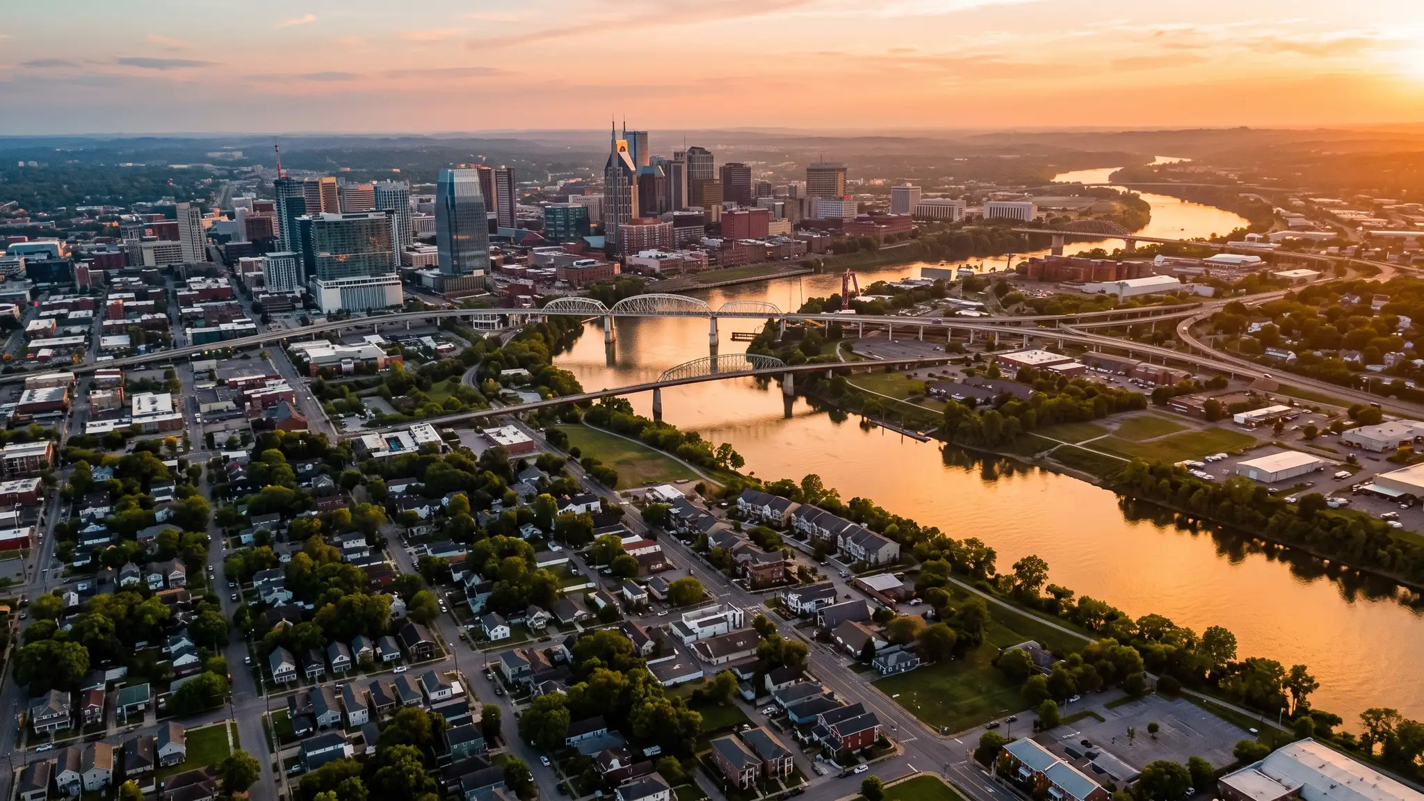 Aerial view of the Cumberland River winding past the Nashville skyline and residential neighborhoods at sunset with golden light on the water